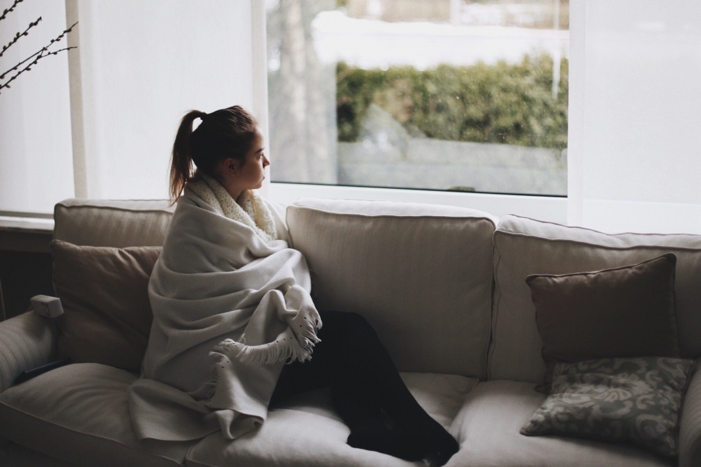 woman with seasonal depression bundled up on the couch looking out the window at the winter sky